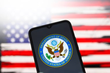ABD Dışişleri Bakanlığı (DOS) logosu bir akıllı telefon ekranında ve arka planda Amerika Birleşik Devletleri bayrağı 3 Aralık 2024 'te Reno, ABD' de bilgisayar ekranında görünür.