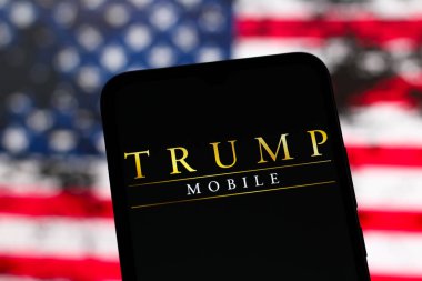 Bu resimde, Trump Mobile logosu akıllı telefon ekranında ve arka planda, Amerika Birleşik Devletleri bayrağı, 17 Haziran 2025, ABD