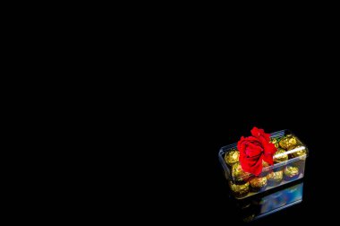 Bir kutu çikolata ve siyah arka planda kırmızı bir gül çiçeği. Bir kutu şeker. Kırmızı gül çiçeği. Altın rengi. Çiçekler ve hediyeler. Sevgililer Günü. Güzellik. Bir kadın için şölen hediyesi..
