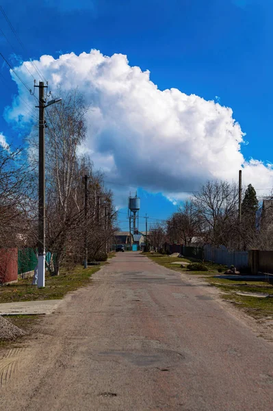 Rural street with a water tower on the horizon. Asphalt road. Water tower. Water supply in the village. Water and metal reservoir. Blue sky. White cumulus clouds. Background image.