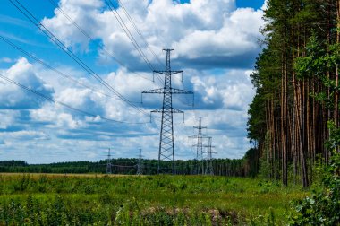 Enerji hatlarının kuleleri doğanın arka planında kablolarla çevrili. Elektrik enerjisinin yüksek voltajlı iletim kulesi. Endüstri ve doğa. Beyaz bulutlu mavi gökyüzü.