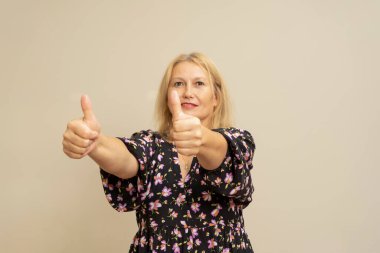 Gülümseyen beyaz kadın, bej arka planda baş parmak gösteriyor.