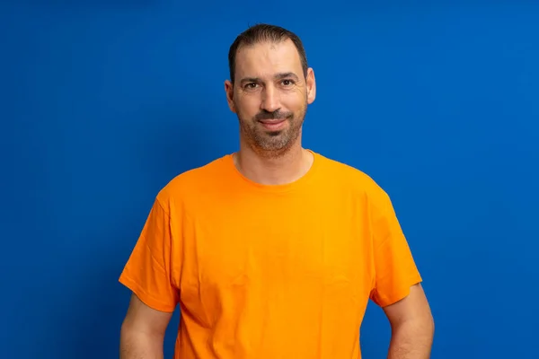 ハンサムな髭の男の孤立したショット カジュアルなオレンジのTシャツを身に着けています 彼の対話者を聞きながら陽気な表情をしています 青を背景にスタジオでポーズ — ストック写真