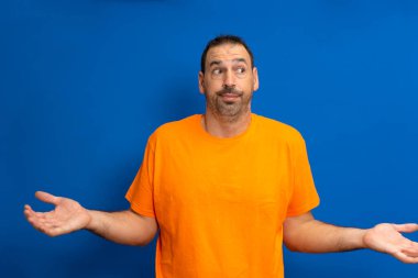 Sakallı, turuncu tişörtlü latin adam, mavi arka planda izole edilmiş, şüphe içinde ve sorgulama jestinde omuz silkiyor..