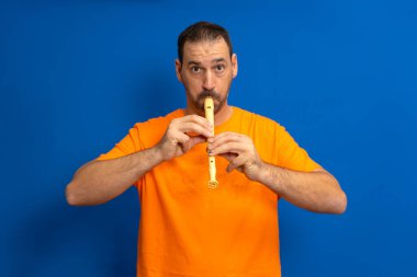 Turuncu tişörtlü İspanyol bir adam mavi arka planda izole bir şekilde okul flütü çalıyor..