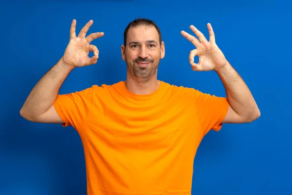 快乐地微笑着 他的惊慌失措的男人留着胡子 穿着一件休闲的橙色T恤 用双手做着一个很好的手势庆祝一件事 被蓝色的工作室背景隔开了 — 图库照片