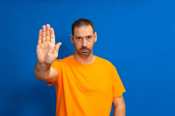 戴着胡子的惊慌男子穿着橙色的T恤 手心紧握着双手 与蓝色的工作室背景隔离 他对现行制度造成的社会和劳工不公正现象感到厌倦 — 图库照片