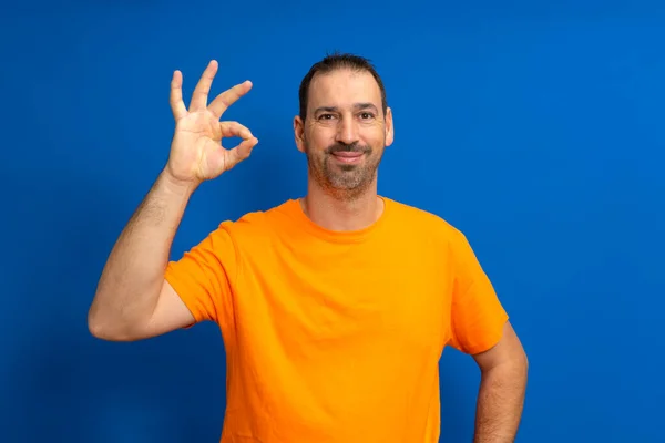 身穿橙色T恤 留着胡子的西班牙裔男人在蓝色的工作室背景下做出了一个很好的手势 — 图库照片