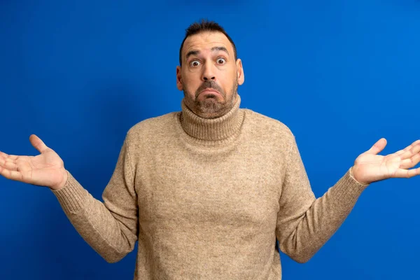 stock image Hesitating bewildered bearded fashionanble man full of doubts, must decide or choose between two proposals, pursed lips, has indecisive expression, shows uncertainty, gestures against blue background