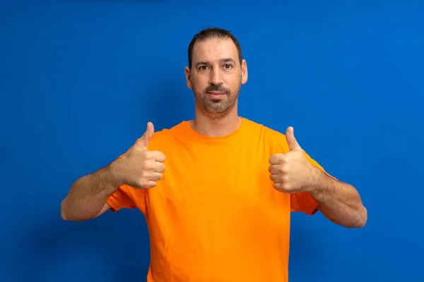 一个喜形于色 留着胡子 身穿橙色T恤的拉丁裔男子的画像 他的大拇指在蓝色背景下被隔离在相机前 — 图库照片