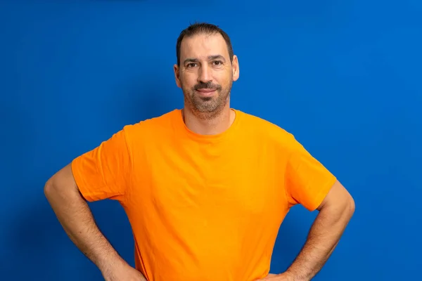 stock image Cheerful bearded man in orange t-shirt posing isolated on blue background, studio portrait. People lifestyle concept. Mock up copy space. Standing with arms akimbo on waist blinking