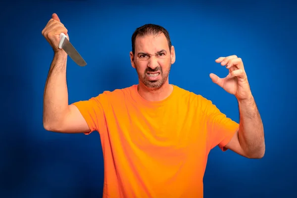 stock image Hispanic man with a beard wearing an orange t-shirt in an aggressive attitude wielding a knife ready to attack, isolated on a blue studio background