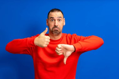 Kırmızı kazaklı, yakışıklı, sakallı, latin bir adamın, mavi arka planda bir aşağı bir yukarı başparmak göstermesi şüpheli.