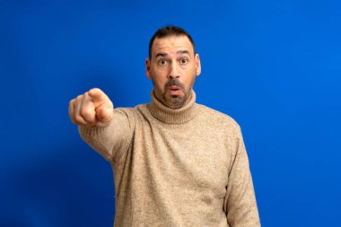 Kahverengi balıkçı yaka kazaklı, sakallı bir İspanyol 'un portresi mavi arka planda izole edilmiş kamerayı işaret ediyor..