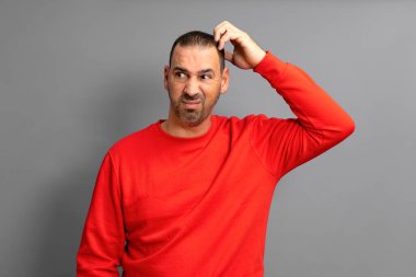 Kırmızı kazaklı, sakallı İspanyol bir adam kaşlarını çatmış ve yüzünü buruşturmuş, mavi arka planda dikiliyor..