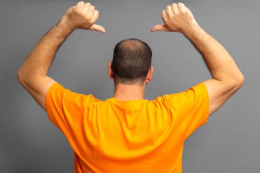 Proud man in orange t-shirt with his back turned pointing with his thumbs at his receding bald spot, isolated on gray background