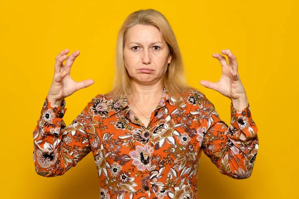 Frustrated blonde woman in print dress with furious expression, raising claws in aggression, experiencing strong emotions. Indoor studio shot isolated on orange background.