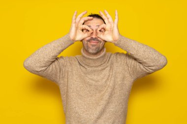 Bearded handsome man wearing turtleneck over isolated yellow background doing ok gesture like binoculars, eyes looking through fingers. Crazy expression