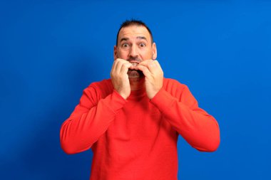 Handsome man wearing red jumper over isolated blue background looking stressed and nervous with hands on mouth biting nails. Anxiety problem.