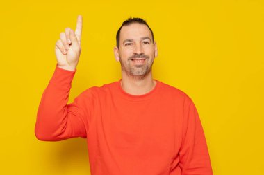 Handsome hispanic man with a beard in a red sweatshirt raising his index finger excited, he has had a great idea and he wants to tell everyone about it. Isolated on yellow background