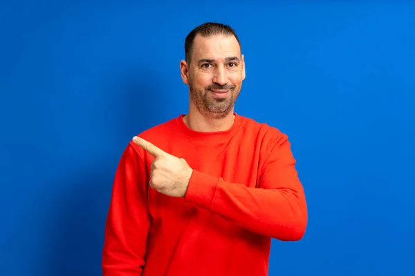 青い背景の上に隔離された側を指している間 カメラに笑みを浮かべて赤いセーターを着て髭を生やしたヒスパニック系の男は 次のパスまたは方向を示す — ストック写真