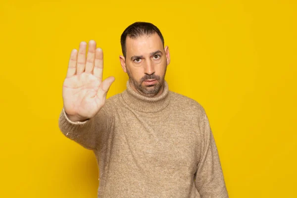 西班牙裔男子 留着胡须 头戴米黄色的套头毛衣 严肃地做出了停止动作 手的手掌与黄色背景隔离在一起 — 图库照片