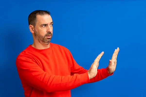 Side view of scared bearded man in beige turtleneck raising hands in sudden fear, showing stop, no, rejection gesture, looking scared, denying problem. Isolated on blue background