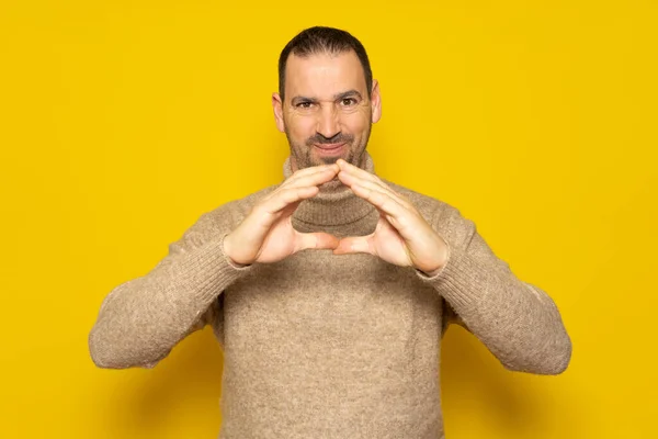 Hispanic man with his hands in a triangle position is giving shape to an evil plan, he smiles maliciously. Isolated on yellow background