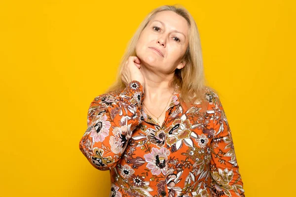 Caucasian blonde woman in a patterned dress scratching her neck with a carefree attitude while hesitating to choose one thing or the other, isolated over yellow background