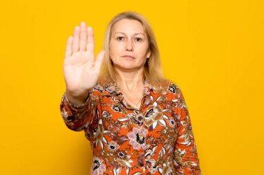 Sarışın, desenli elbiseli, avucunu dur işaretinde kaldıran, sarı arka planda izole bir beyaz kadın.