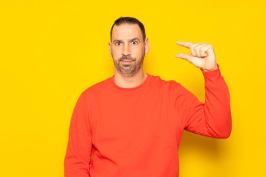Sakallı yakışıklı İspanyol bir adam. Elinde kırmızı bir süveter, parmakları ve kamerasıyla küçük bir işaret yapıyor. Ölçüm kavramı. Sarı arkaplanda izole edilmiş.