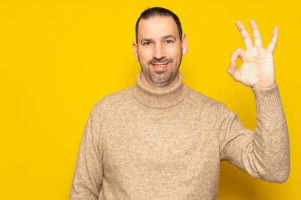 stock image Bearded hispanic man in his 40s wearing a beige turtleneck making the ok gesture while smiling at the camera, isolated over yellow background