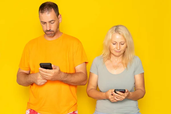 Middle aged couple looking at their cell phone ignoring each other isolated on yellow studio background. Concept of social distancing due to technology