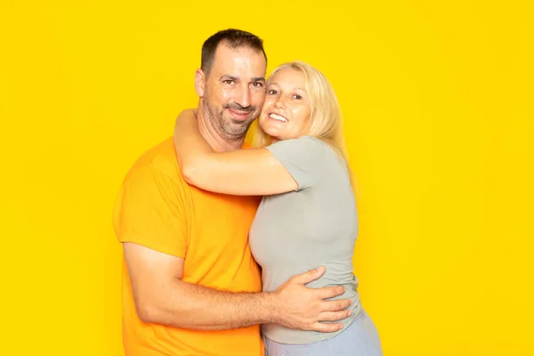 Real Caucasian couple in their 40s tenderly hugging each other demonstrating their love and commitment, isolated on yellow background