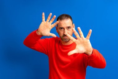 40 'larında sakallı, koruma modunda elleri açık, mavi stüdyo arka planında izole edilmiş İspanyol bir adam.