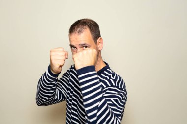 40 'larında sakallı İspanyol bir adam, çizgili bir süveter giyiyor, yumrukları savunma pozisyonunda dövüşmeye hazır, bej rengi izole bir arka planda izole edilmiş.