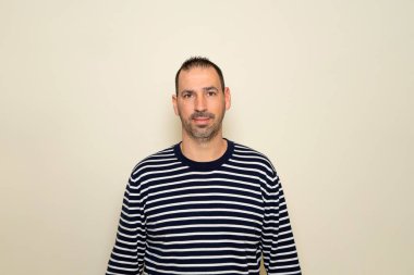 Sakallı, çizgili süveter giyen bej renkli stüdyo arka planında gülümseyen İspanyol bir adam.
