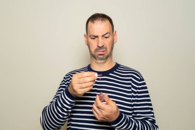 40 yaşlarında, ispanyol sakallı, çizgili bir süveter giyen, tiksindirici ve şaşkın bir yüzü olan, bej renkli stüdyo arka planında izole edilmiş bir tırnak cilasıyla uğraşan bir adam.