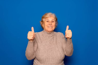 70 yaşlarında, koyu bej süveter giyen, pozitiflik ve memnuniyet belirtileri gösteren, mavi stüdyo arka planında izole edilmiş beyaz bir kadın.