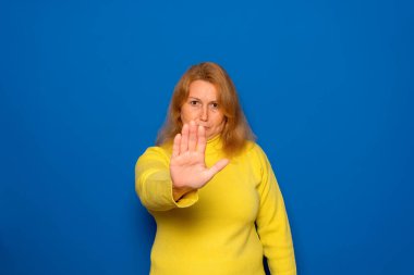 40 'larında beyaz bir kadın kameraya ciddi bir ifadeyle bakıyor. Sağ elini kaldırdı, avuç içi ileri, açık bir duruş hareketi yaptı. Mavi arkaplanda izole