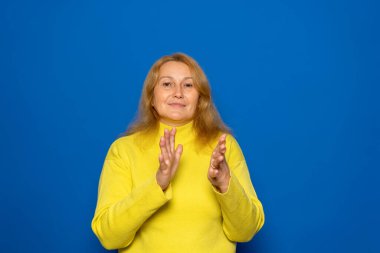 40 'lı yaşlarda beyaz bir kadın kendini mutlu ve başarılı hissediyor. Gülümsüyor ve alkışlıyor. Mavi duvara el çırparak tebrik ediyor.