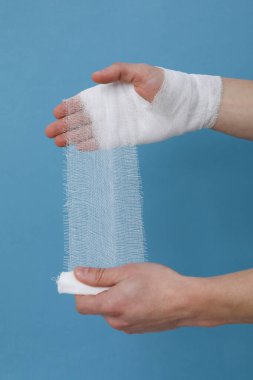 Concept of help during an injury, man wrapping hand in bandage on blue background