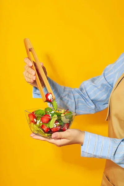 Woman Holds Salad Strawberry Wooden Tongs Yellow Background — Φωτογραφία Αρχείου