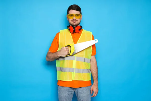 Worker with saw in goggles and protective ear muffs on blue background