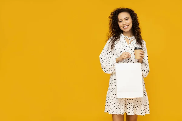 Young woman with shop bag and coffee on yellow background