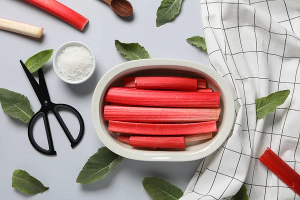 stock image Rhubarb stalk in bowl, mint, salt, scissors and white checkered towel on gray background, flat lay