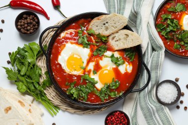 Beyaz arka planda Shakshuka, havlu, baharat, kırmızı biber ve pide olan tavalar.