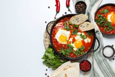 Beyaz arka planda Shakshuka, havlu, baharat, kırmızı biber ve pide olan tavalar, metin için yer