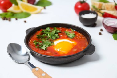 Shakshuka, kaşık, ekmek dilimleri, domates, limon, fesleğen, soğan ve baharatlar beyaz arka planda, kapat