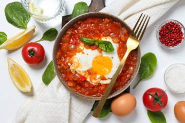 Shakshuka güvertede, çatal, havlu ve malzemeler beyaz arka planda.